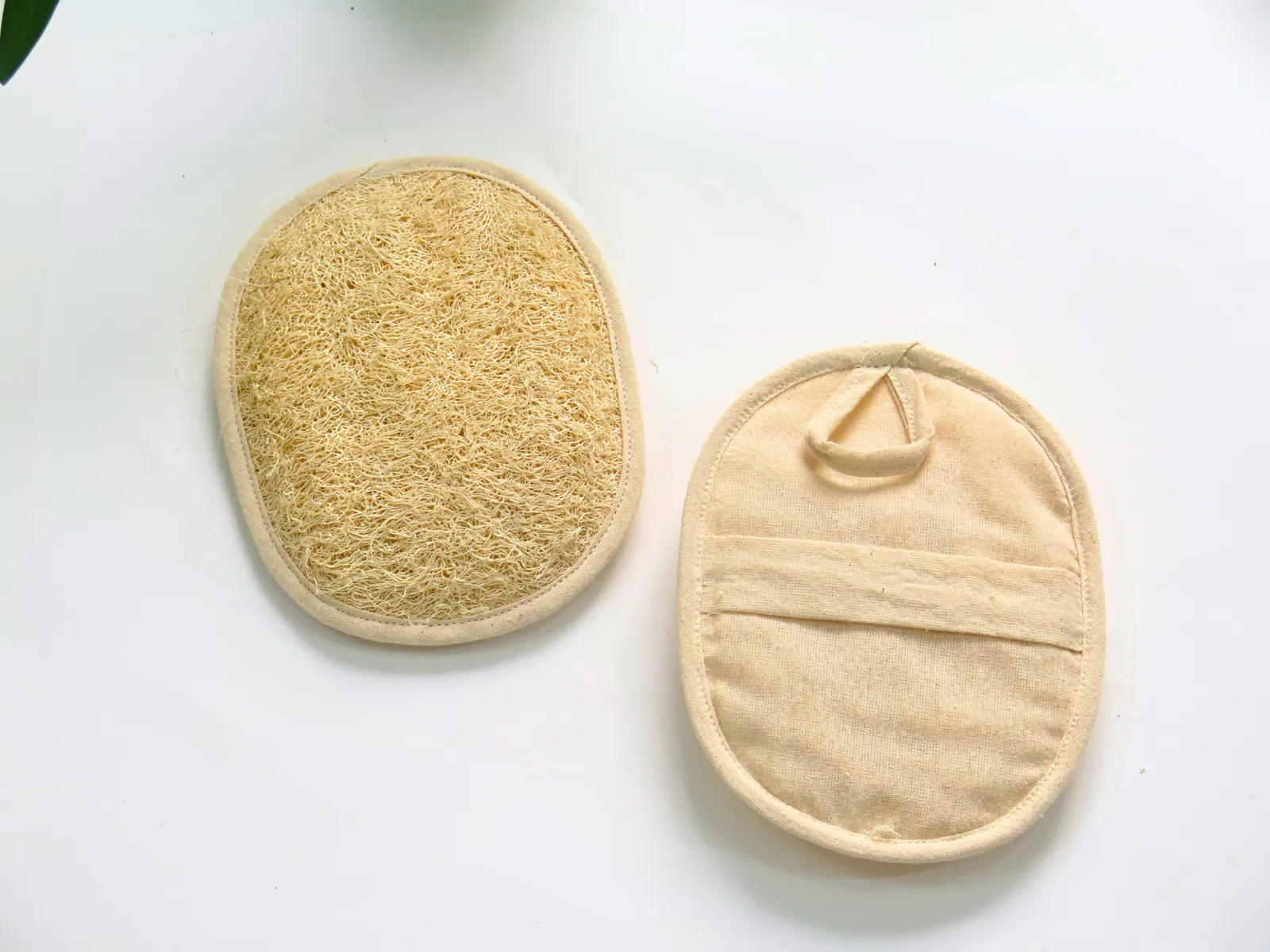 Natural loofah oval pad beside its fabric-backed side with a hand strap on a white surface
